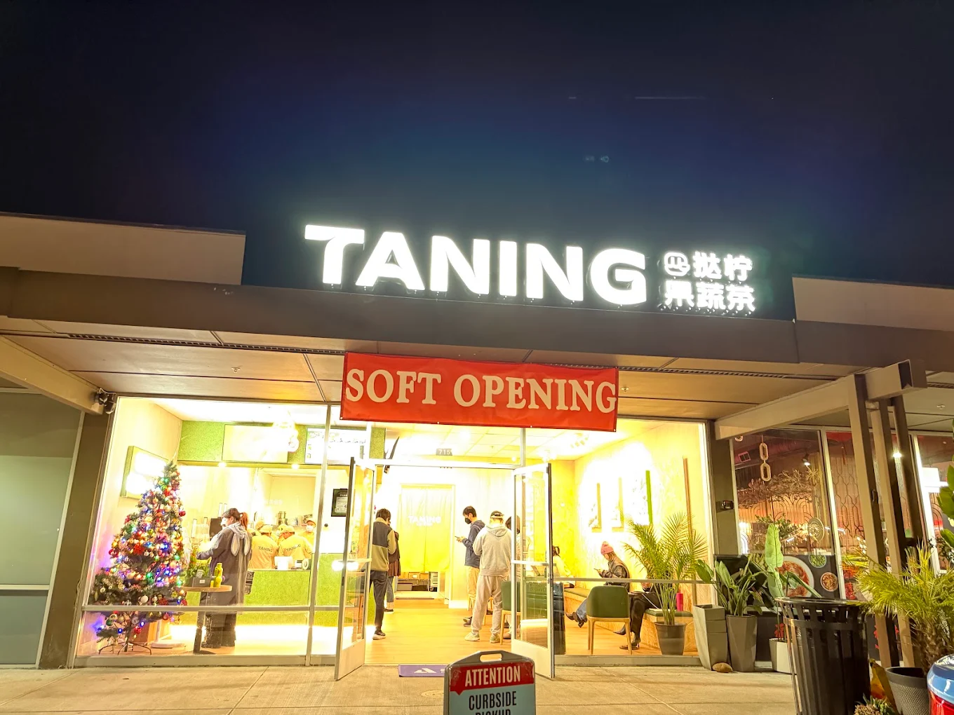 TANING store exterior at night with a red "SOFT OPENING" banner and customers visible inside the lit shop.